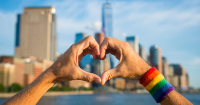 Hands in heart formation with rainbow armband and NYC skyline in the background (Photo Credit: shutterstock)