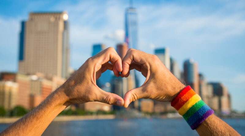 Hands in heart formation with rainbow armband and NYC skyline in the background (Photo Credit: shutterstock)
