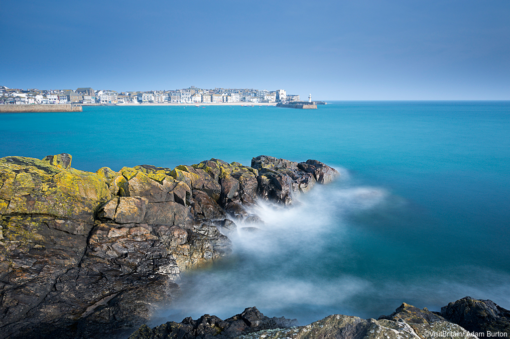 View across calm turquoise sea water to St Ives town and harbour. (Photo Credit: ©VisitBritain/ Adam Burton)