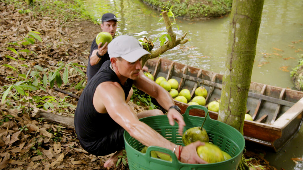 In the Thailand countryside, Ian and Joe fill their canoe full of pomelo fruit found in a river and transport it to land to fill five baskets. (Photo Credit: CBS)